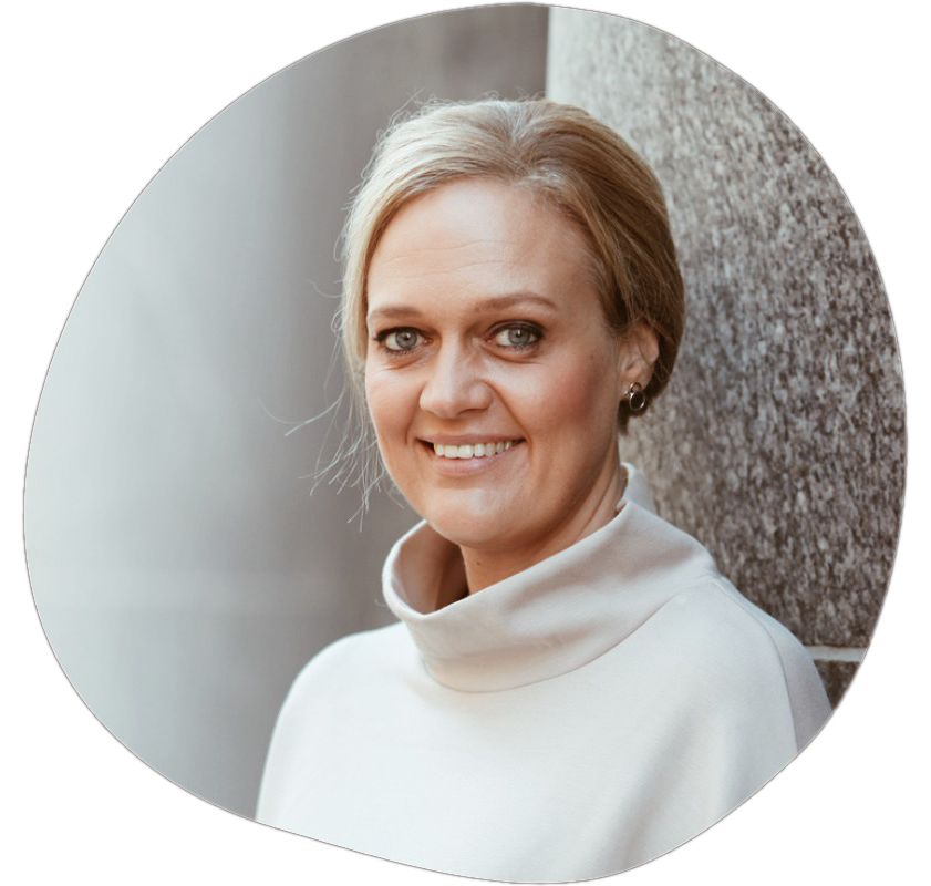 Porträtt av en leende kvinna med ljusbrunt hår, iförd en vit tröja, stående mot en betongvägg.