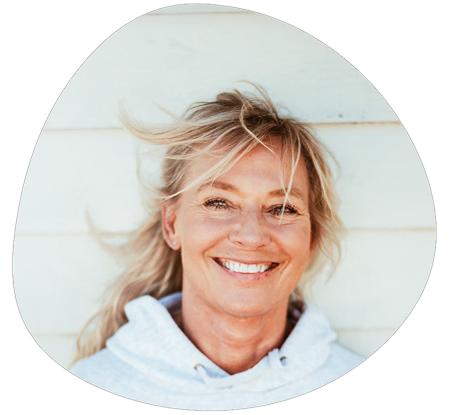 En kvinna med blont hår ler mot kameran, bär en grå hoodie, står framför en vit vägg.