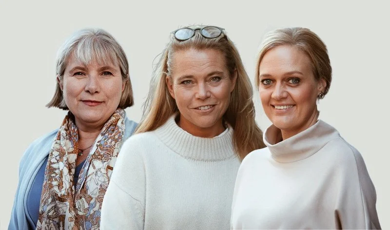 Tre kvinnor står nära varandra och ler mot kameran, mot en ljus bakgrund.