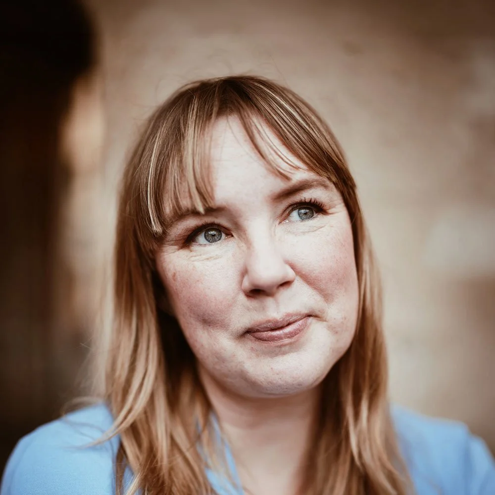 En man med rakhår, glasögon och nose ring, som ler lätt, bär en mönstrad skjorta, står i ett rum med bokhylla i bakgrunden.