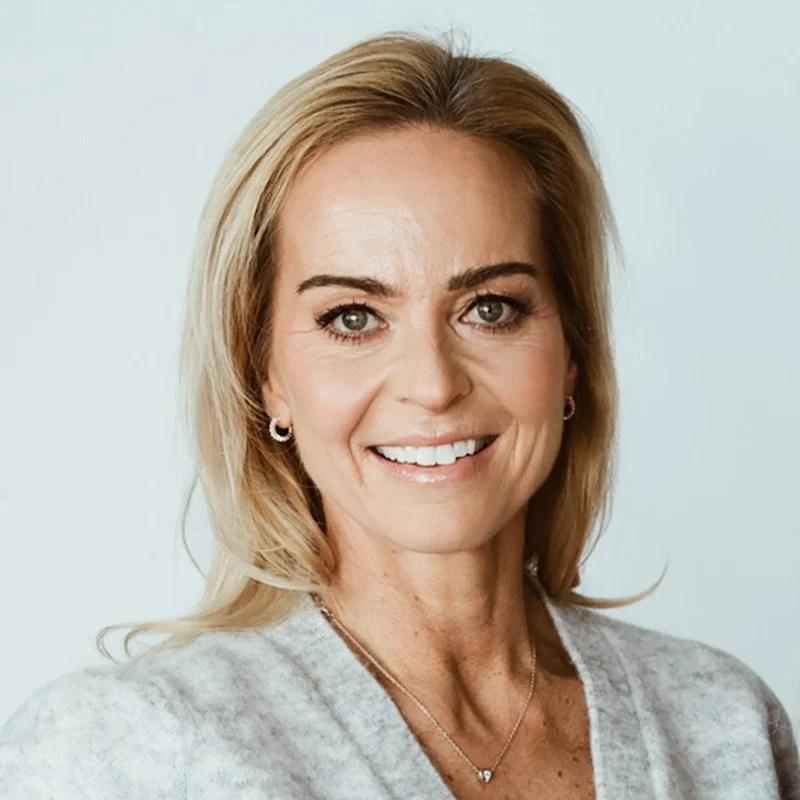 Porträtt av en leende kvinna med blond hår, silver örhängen och en silver halskedja, klädd i en ljusgrå tröja, mot en vit bakgrund.