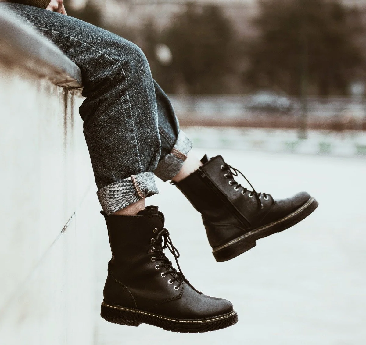 Person som sitter med benen utsträckta, bär svarta läderstövlar och rullade jeans, mot en beigefärgad vägg, utomhus.