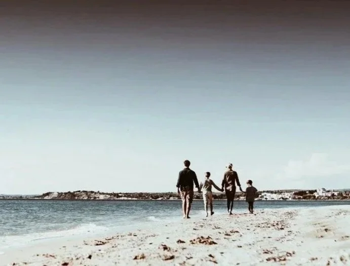 En familj med två vuxna och två barn som går hand i hand på stranden.