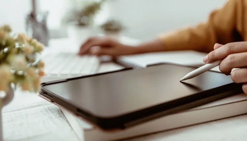 Person som använder en surfplatta med en stylus, på ett skrivbord med blommor och en dator i bakgrunden.