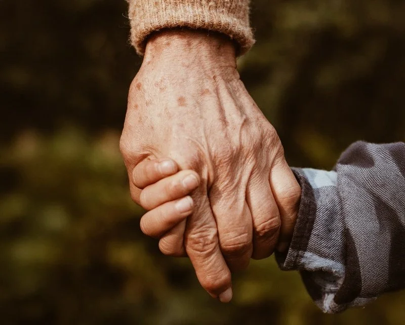 Två personer håller varandra i handen, en äldre och en yngre, utomhus med natursbakgrund.