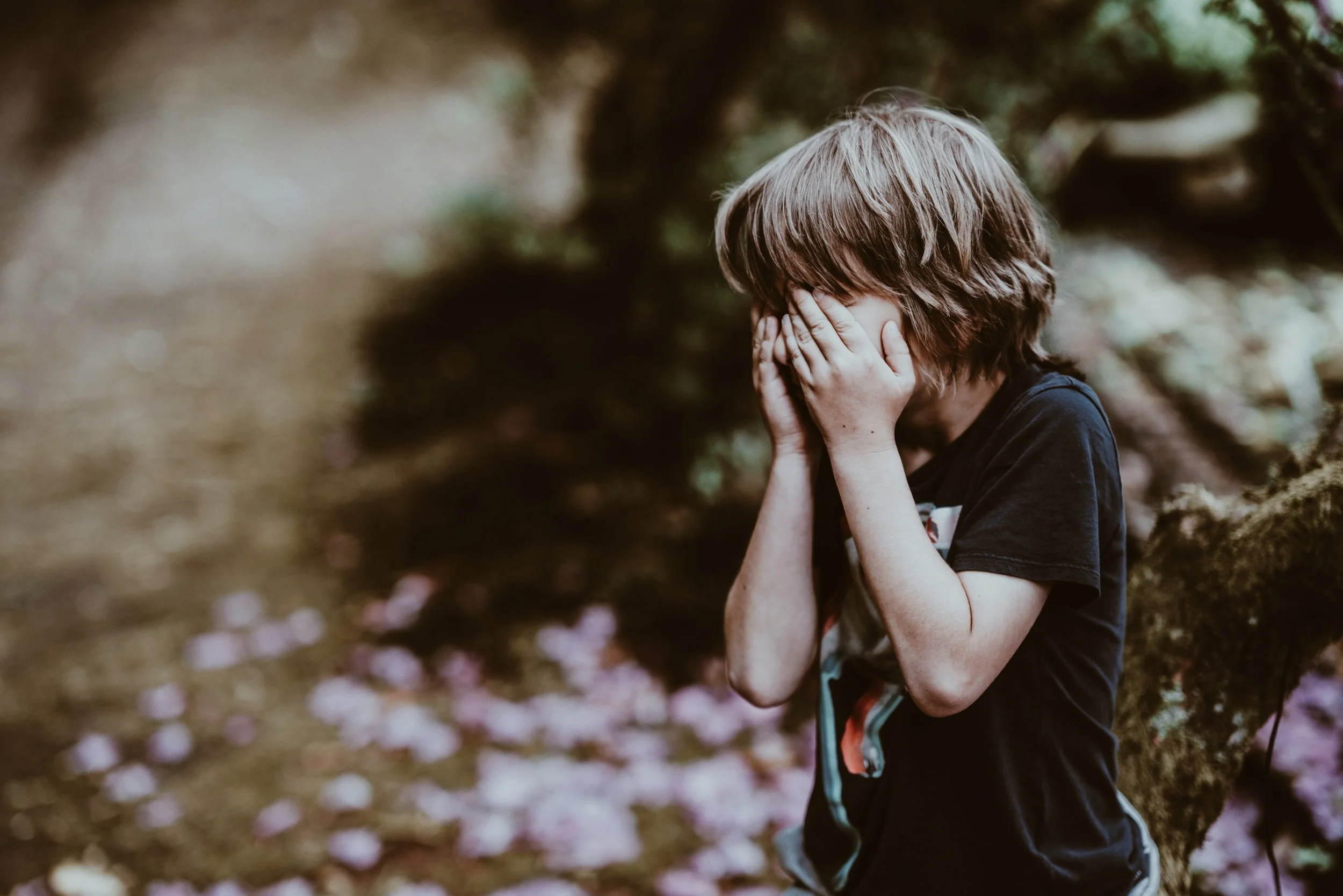 En ung pojke med långt hår som täcker ansiktet med händerna, sitter i en skog med blommor på marken och ett träd bakom honom.