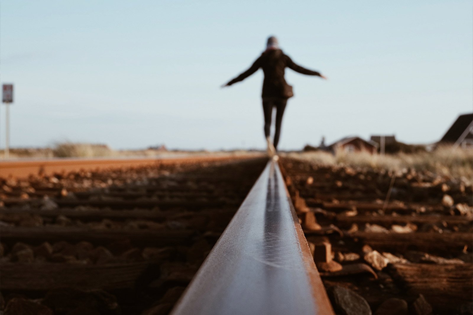 En person balanserar med armarna utsträckta på ett spår av ett järnvägsspår, med utsikt mot horisonten under en ljus himmel.