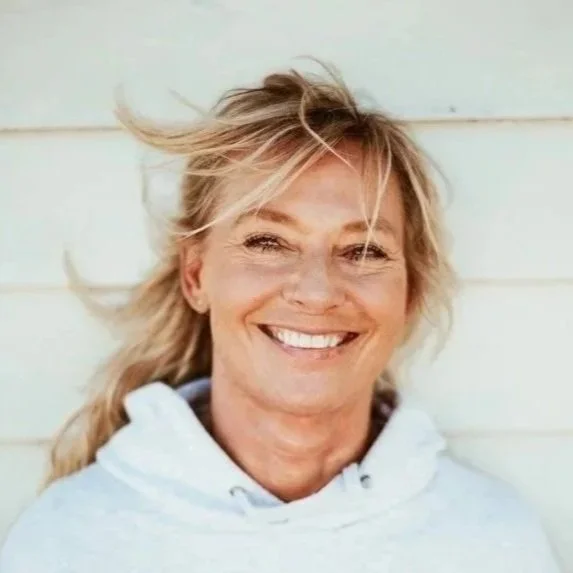 En skrattande kvinna med blont hår som blåser i vinden, iklädd vit hoodie, stående framför en ljus vägg.