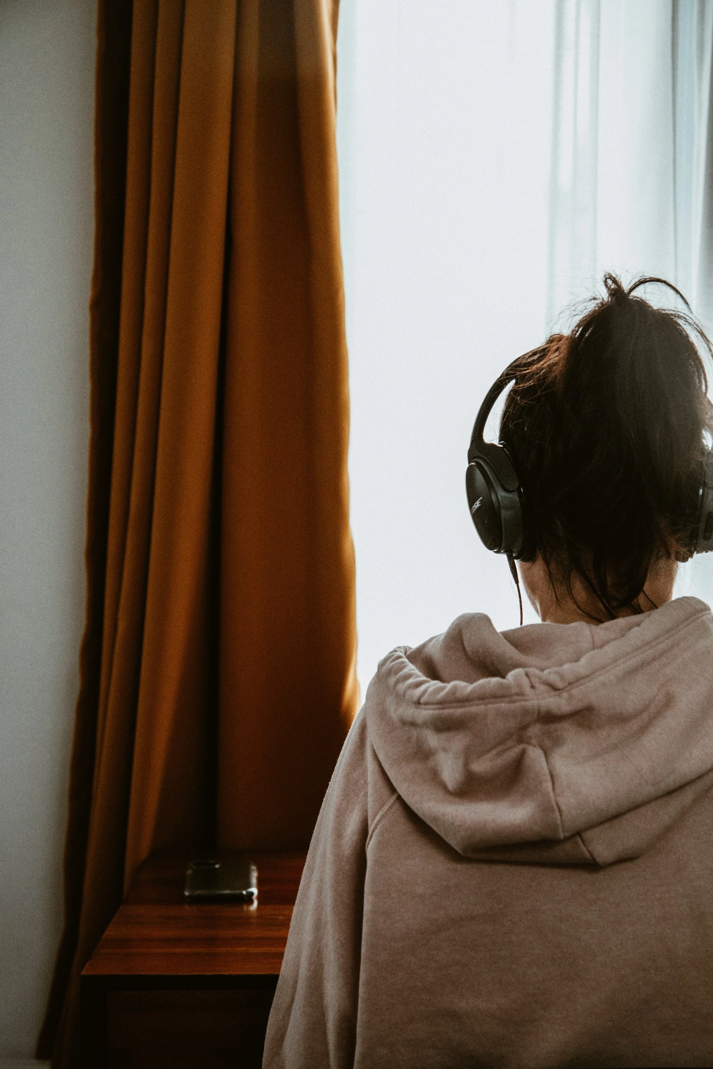 Person med mörkt hår och hörlurar tittar mot ett fönster, klädd i en beige hoodie, nära en guldfärgad gardin och ett trähängbord.