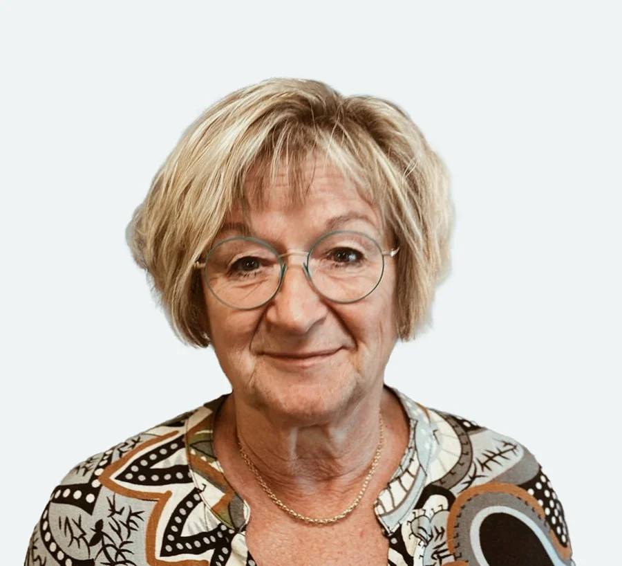 En äldre kvinna med kort, ljust hår och glasögon, ler mot kameran, bär en mönstrad blus