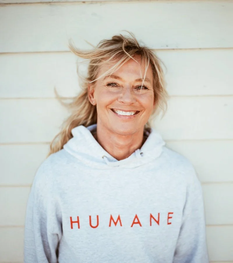 En leende kvinna med blont hår i en grå hoodie med texten 'HUMANE' står framför en vit vägg.