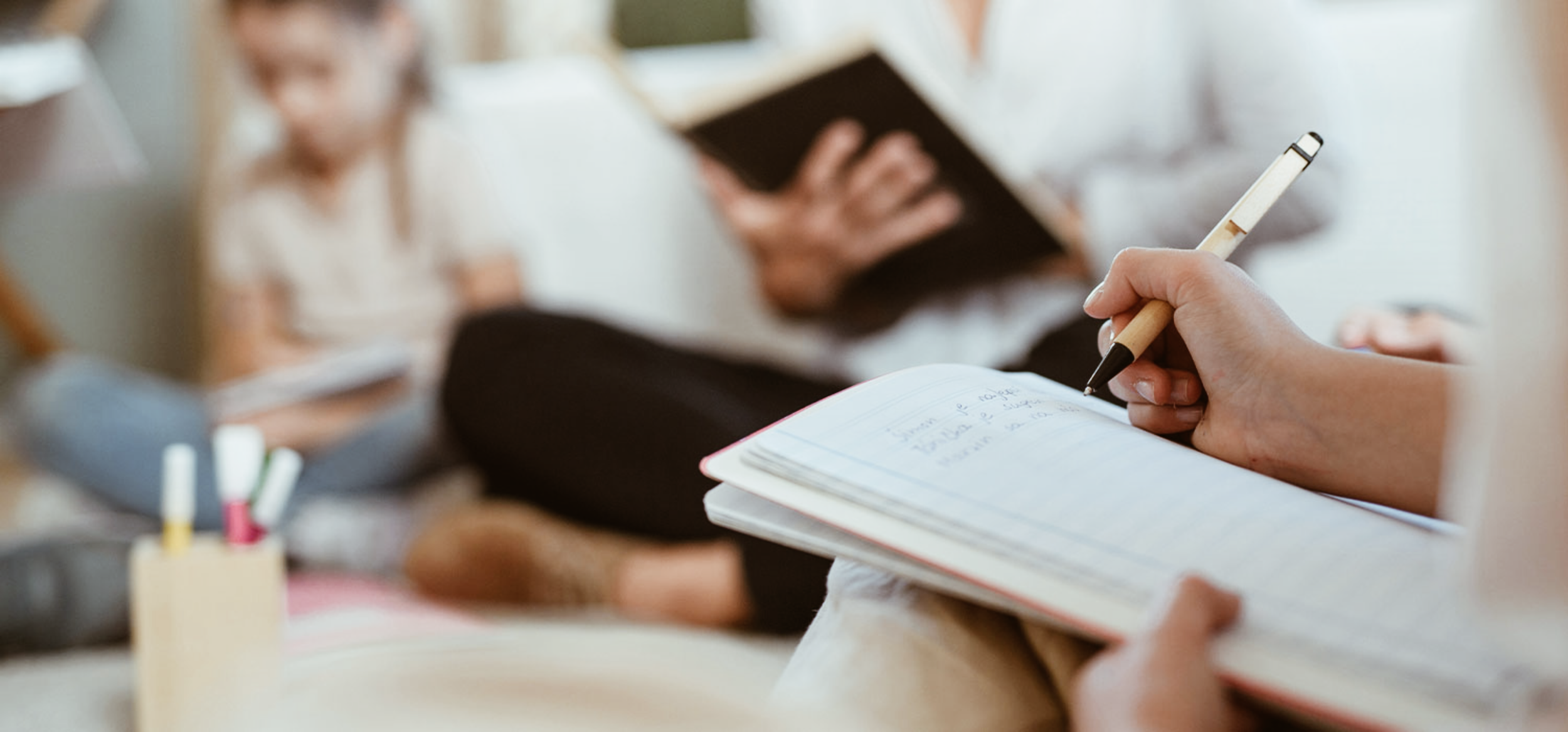 En person som skriver i en öppen dagbok med en penna, medan två andra personer sitter i bakgrunden, en med ett bokhögtal och en annan person som håller i en bok.