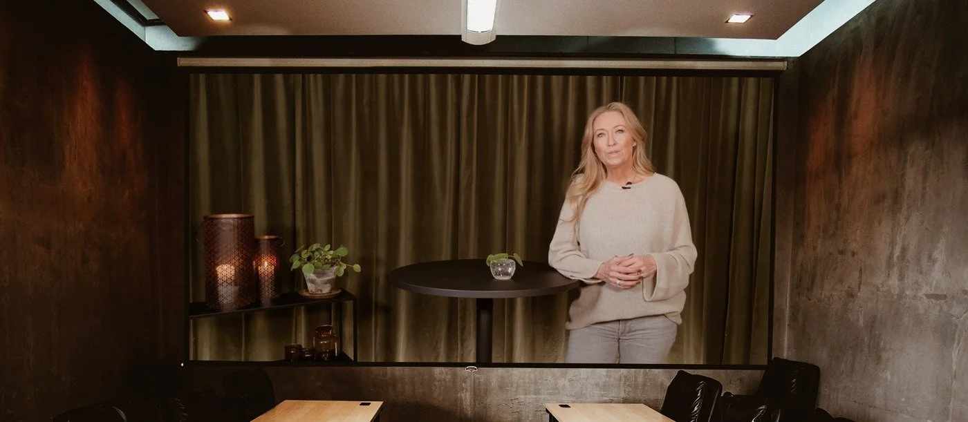 En woman står framför en filmduk i ett rum, som förbereder sig för att prata eller visa något, med en rund svart bord och dekorativa föremål på en hylla framför duken.