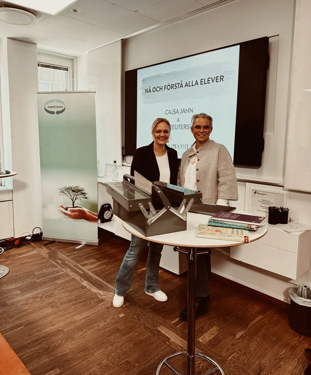 Två kvinnor står tillsammans framför en presentation i ett klassrum, med en projektor och en tavla som visar texten "NÄ OCH FÖRSTÅ ALLA ELEVER". En av kvinnorna har på sig en svart kavaj och den andra en ljus kappa. Det finns en rund bord med vissa böcker och ett arkiv. En banner med en träd omgiven av moln är i bakgrunden.