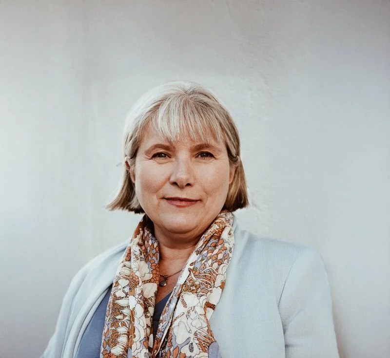 En medelålders kvinna med kort, grått hår, iklädd ljus blazer och mönstrad scarf, står mot en neutral bakgrund.