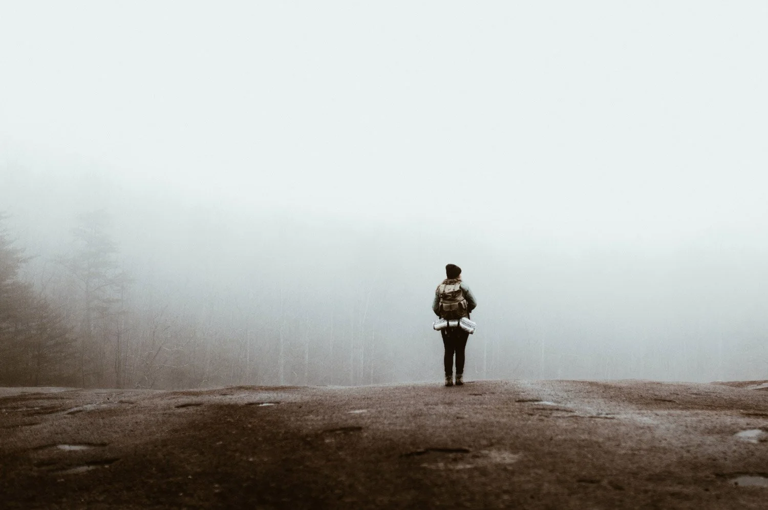En person med ryggsäck som står på en oklar väg och tittar ut mot en tät dimma i en skoglig miljö.