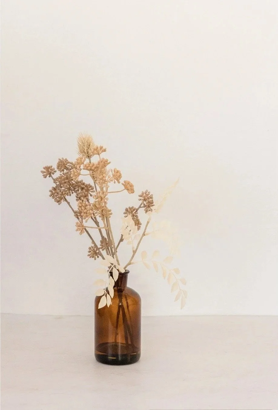 En brun glasvas med torkade blommor och växter på en vit yta.