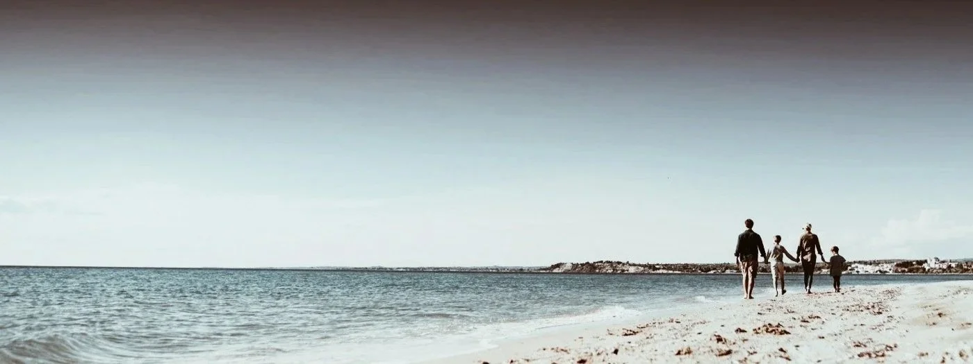 En familj med två vuxna och två barn promenerar på stranden mot horisonten.