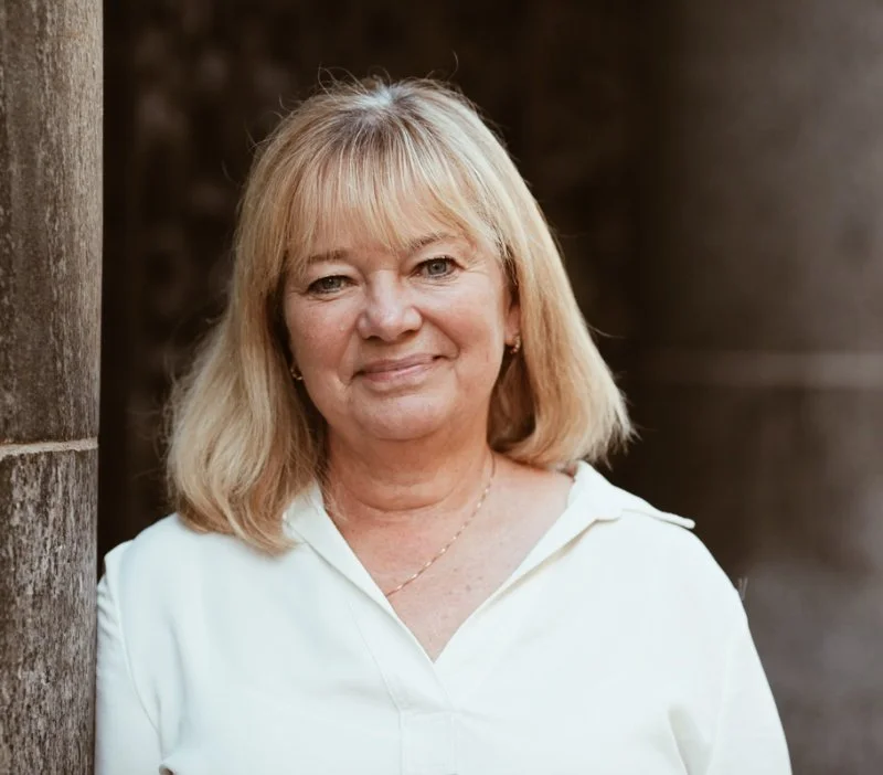 En äldre kvinna med blont hår, klädd i vit skjorta, står utomhus framför en stenvägg och ler.