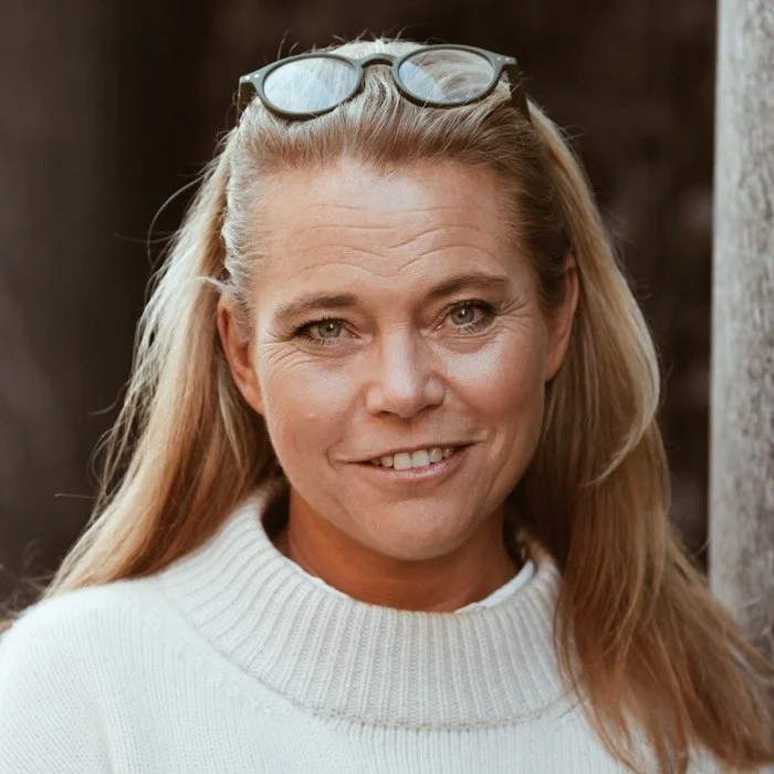 En kvinna med lång blont hår, som bär solglasögon på huvudet och en vit tröja, utomhus.