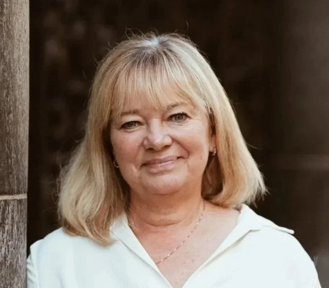 En medelålders kvinna med blont hår och vit topp, står utomhus vid en trästolpe.