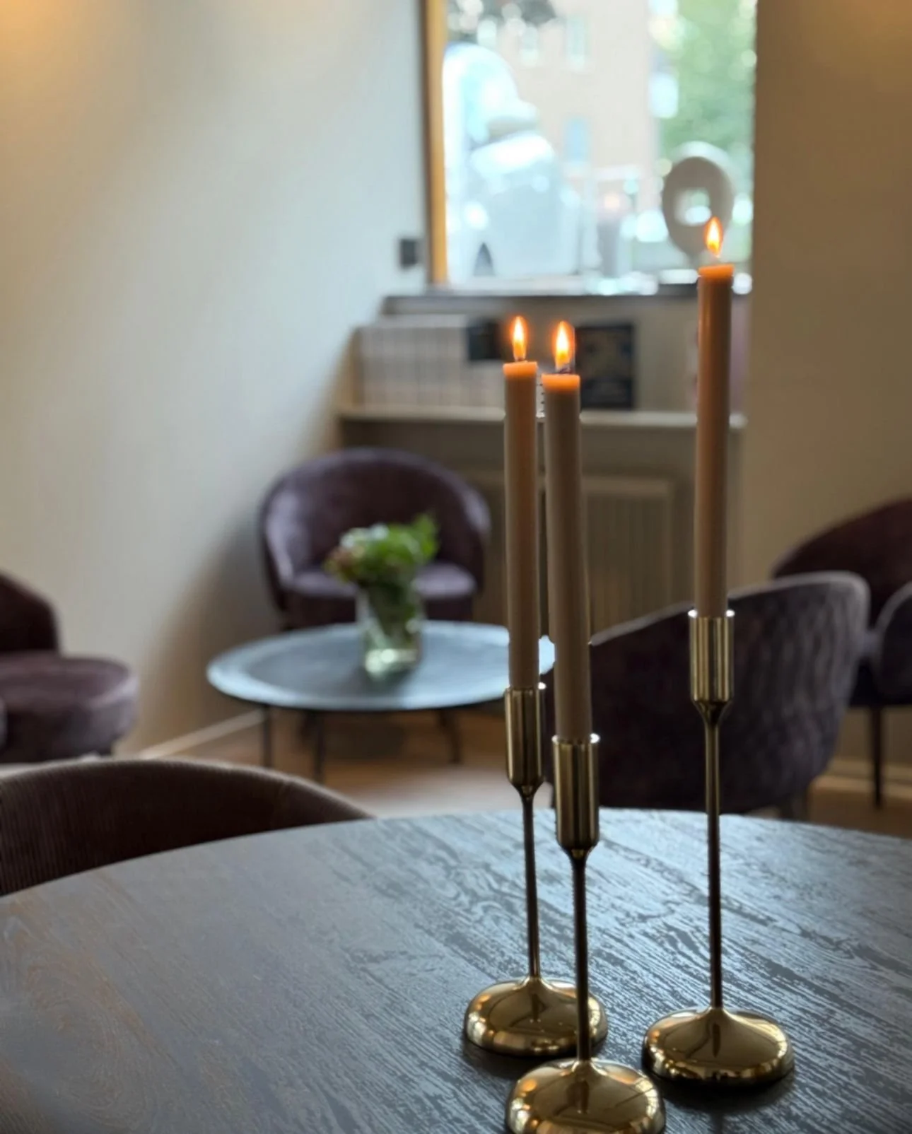 Tända ljus på ett bord i ett inomhusrum med stolar och ett fönster i bakgrunden.