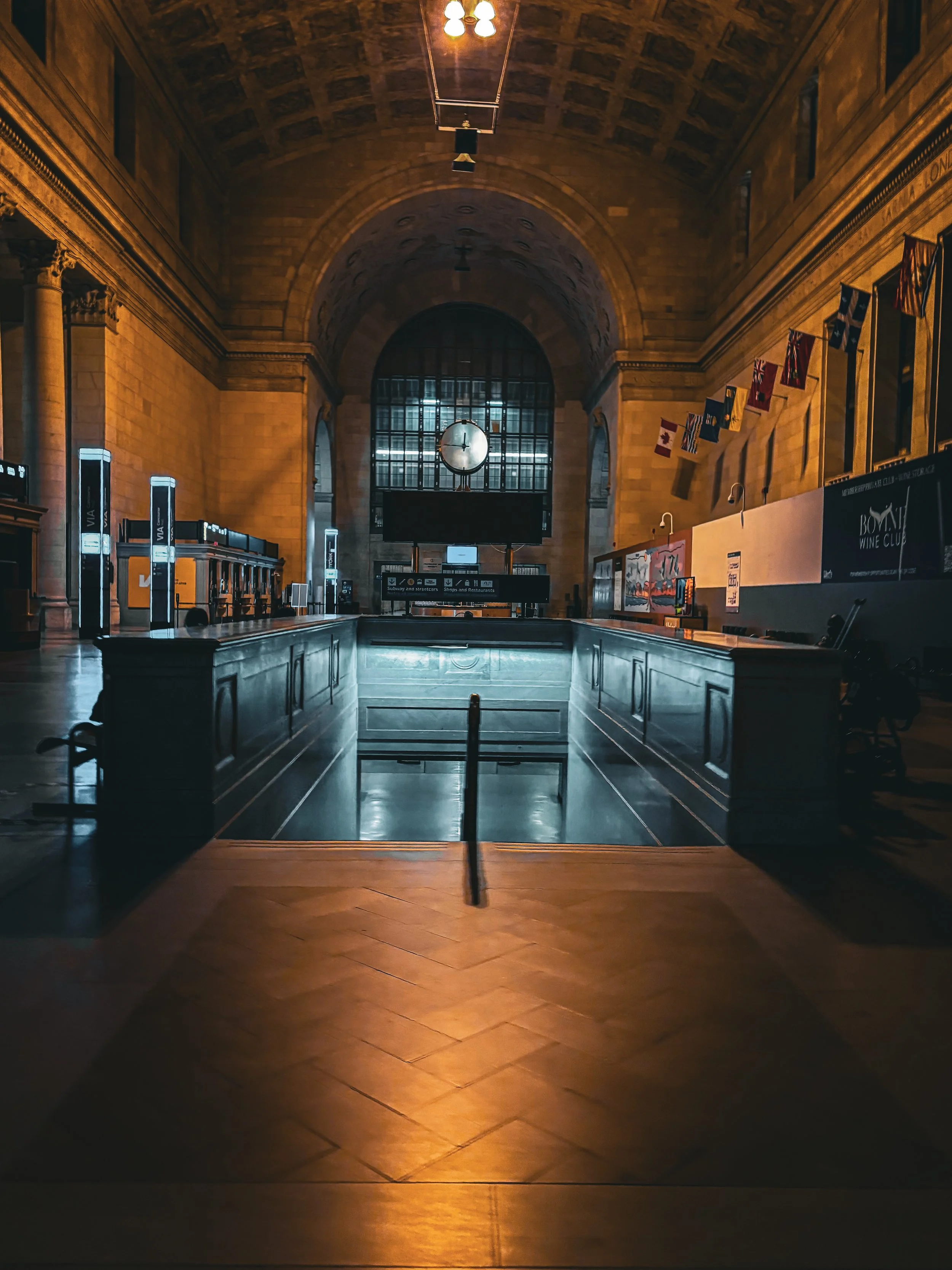 Interior of a grand train station with arched ceiling, large clock, and row of colorful flags. The space is dimly lit, highlighting architectural details and a center staircase.
