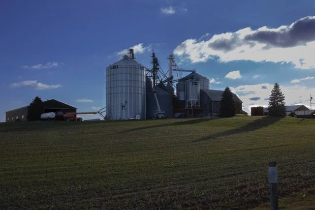 Farm structures with silos and machinery on a grassy field under a partly cloudy sky.