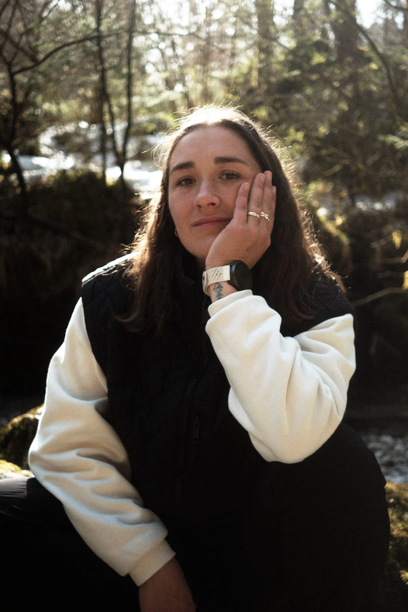 Junge Frau in einem Wald sitzt am Fluss, trägt eine schwarze Weste und ein weißes Sweatshirt, während sie die Hand an ihr Gesicht legt. Jenny Schweiger, Schenewa, Emergenz, Erik Schereder