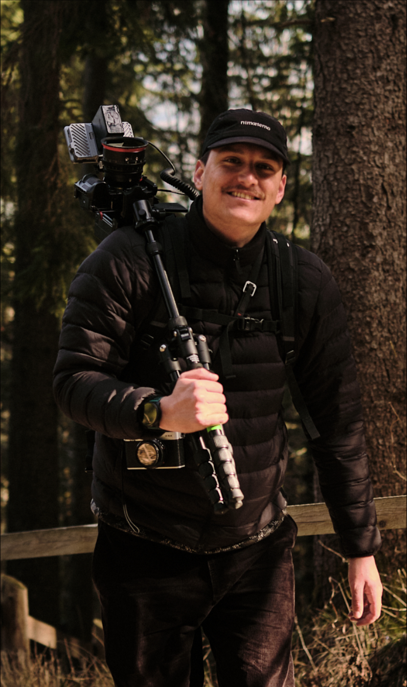 Mann steht in einem Wald mit Rucksack und Kameraausrüstung, trägt schwarze Jacke und Mütze, lächelt. Erik Schereder