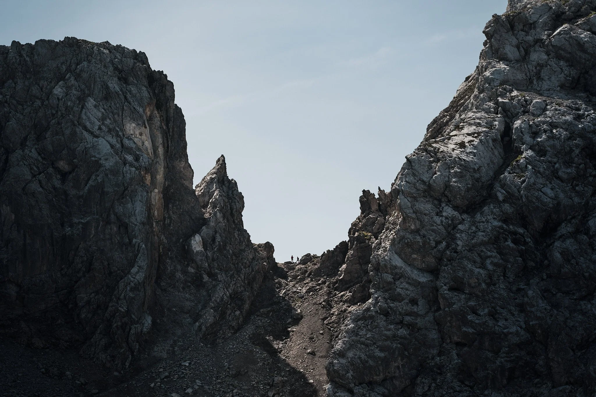 Zwei Wanderer auf einem felsigen Bergpfad zwischen großen, dunklen Felsen unter blauem Himmel. Johannes Rapprich