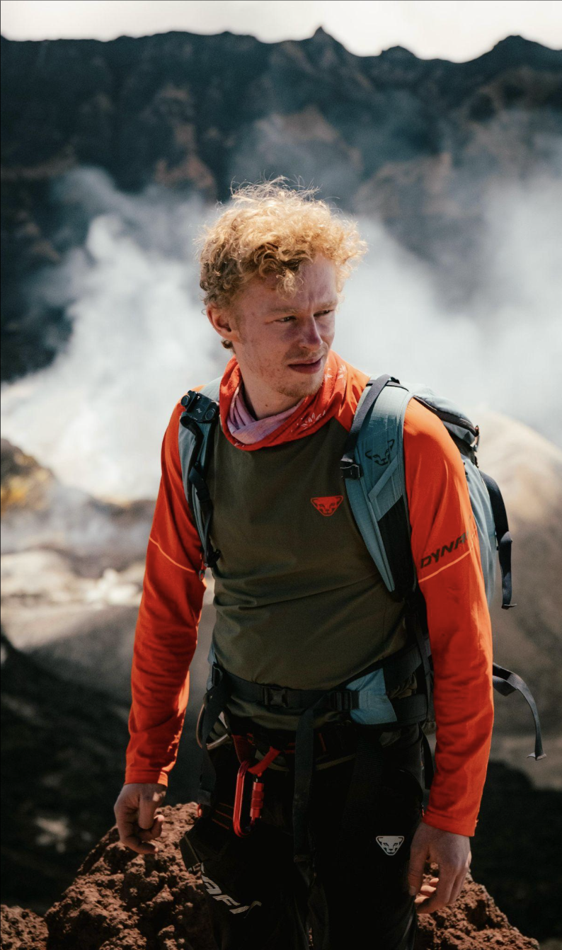 Junger Mann mit Rucksack auf einer felsigen Klippe, im Hintergrund Wasserfall und Berge. Vincent Götte