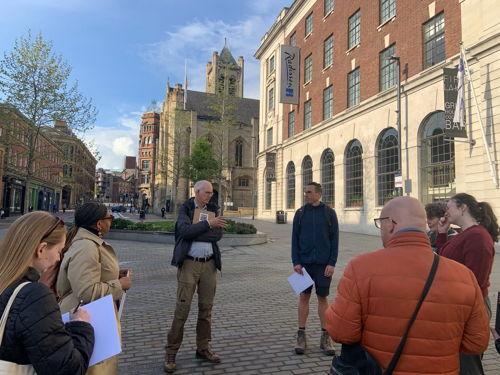A group of people gathered outdoors in an urban setting, listening to a man speaking while holding papers. The background features historic and modern buildings, including a church and a red brick building, under a blue sky with some clouds.