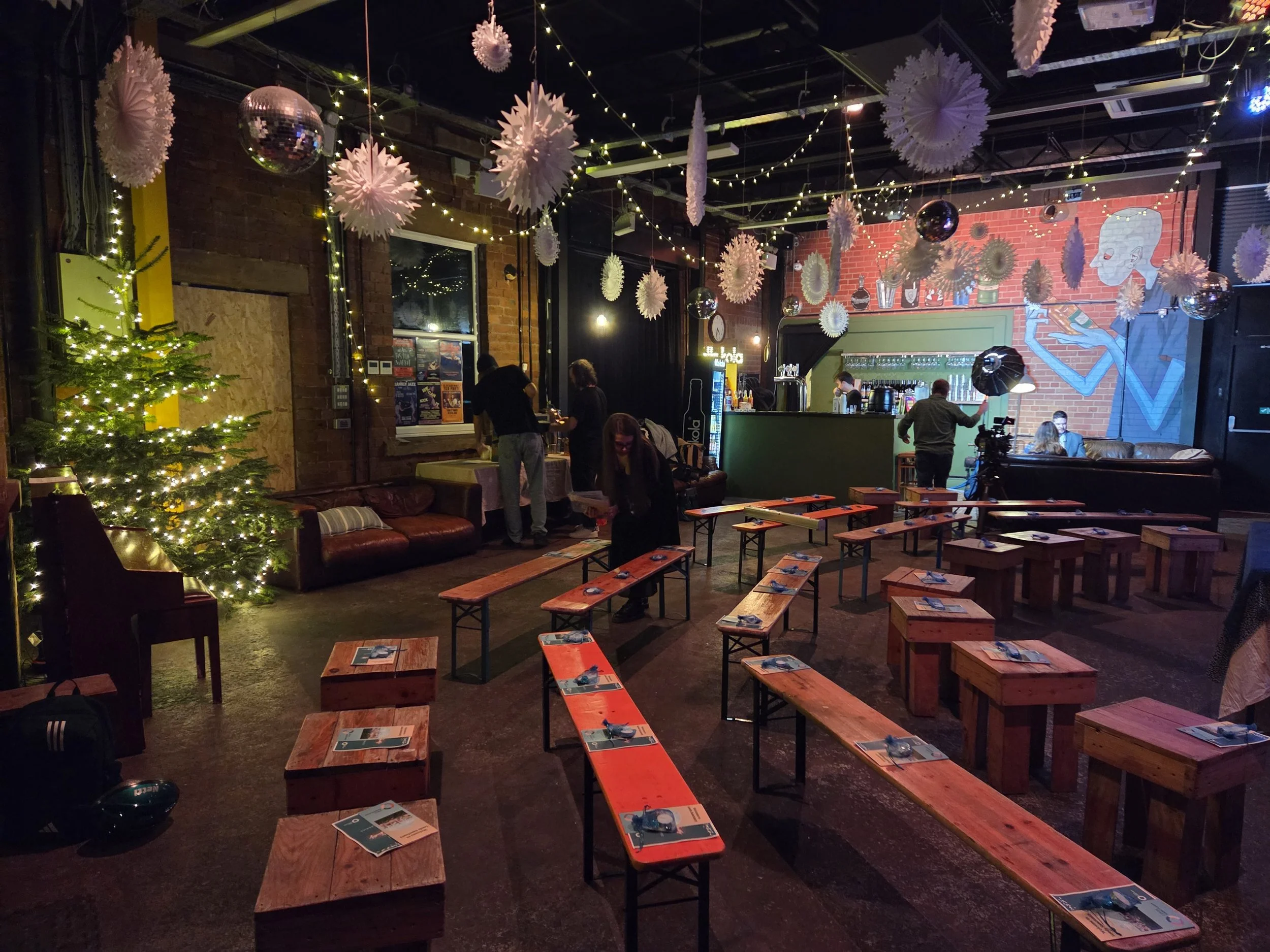 Indoor venue decorated for a festive event with string lights, hanging paper decorations, a Christmas tree with lights, and tables with place settings. People are preparing for our Blue Christmas event near a bar area with a mural on the wall.