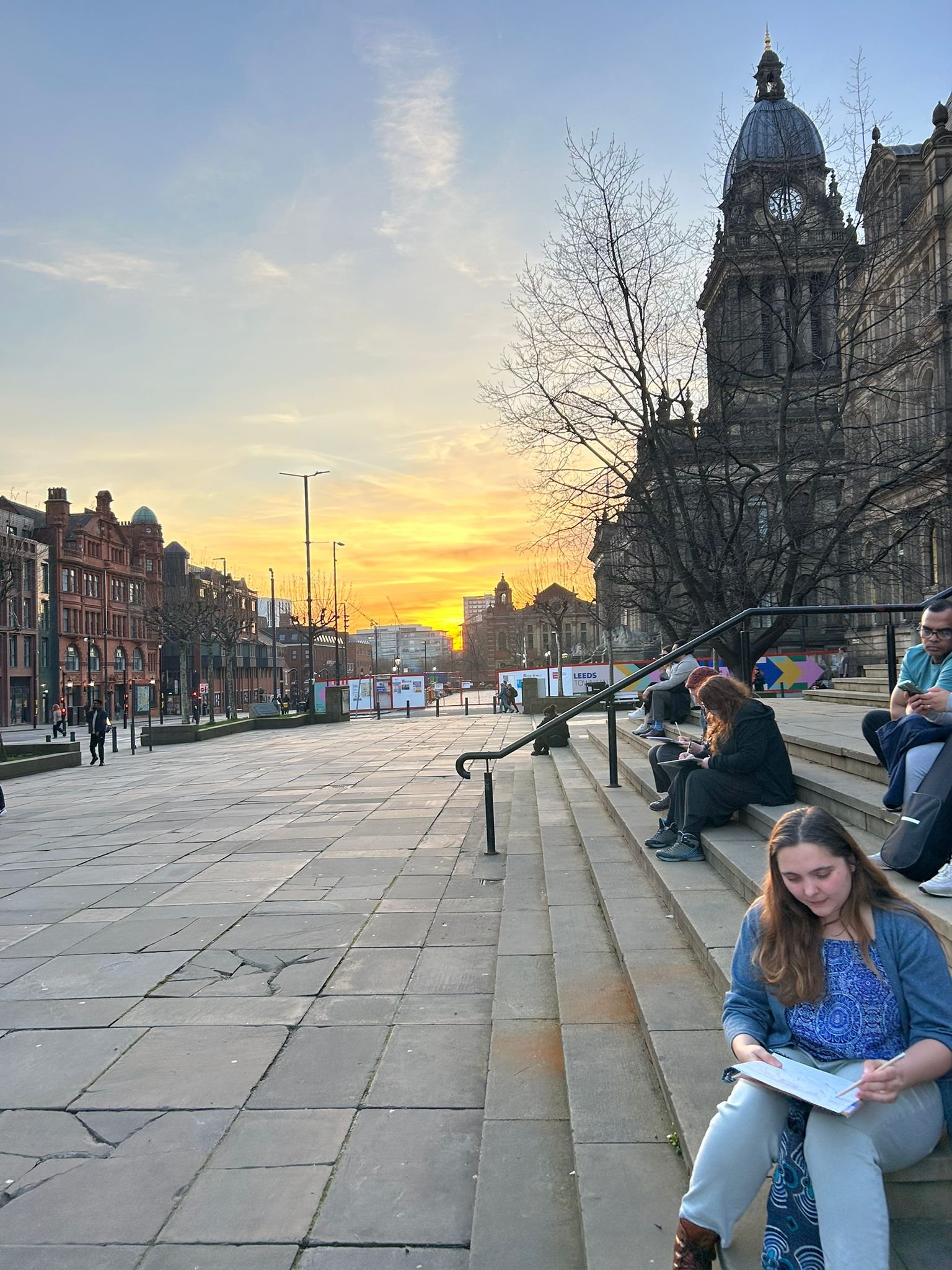 People sitting on steps at sunset in an urban area with historic buildings and a church clock tower in the background.