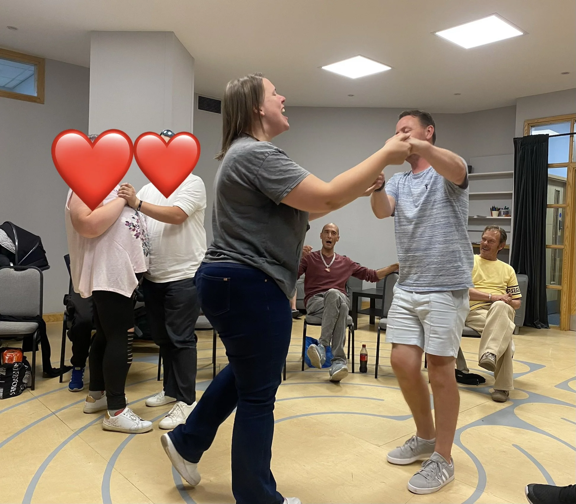 Two people are dancing closely, holding hands and smiling in a room with several seated witnesses. One person's face is partially obscured by two red heart emojis. The room has high ceilings, windows, and wooden accents, with spectators seated and watching the dance.