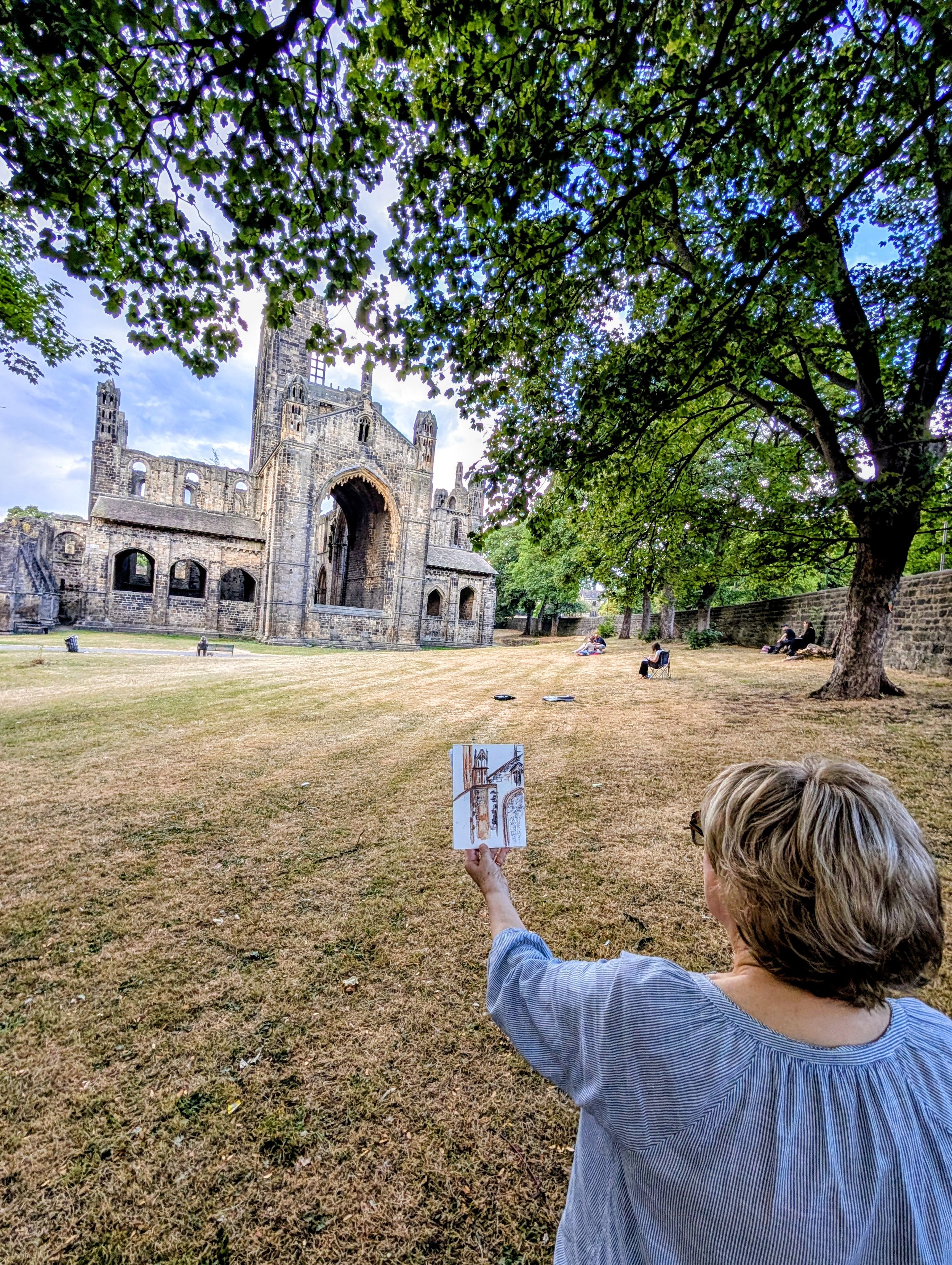 A woman sitting in on the grass in the grounds of Kirkstall Abbey, holding a sketchbook with an illustration of the building.