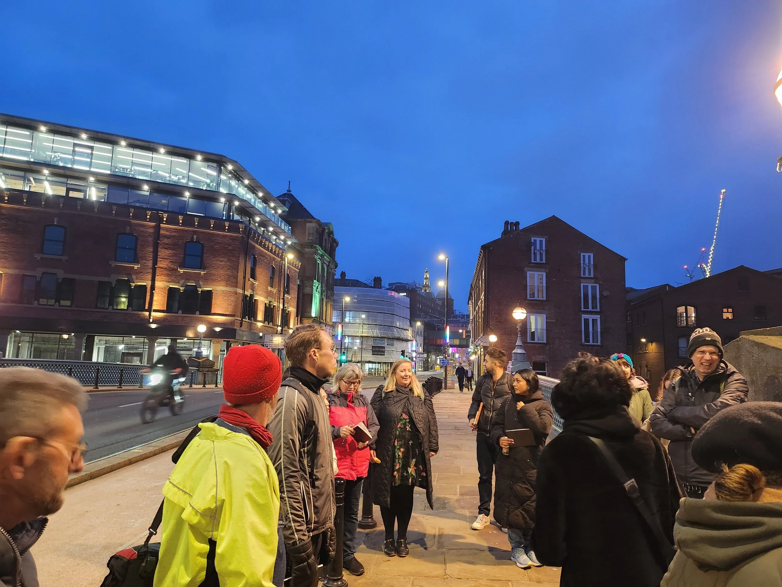 Crowd of people gathered outside on a city street in the evening, with buildings and streetlights illuminated, and a clear blue sky overhead.