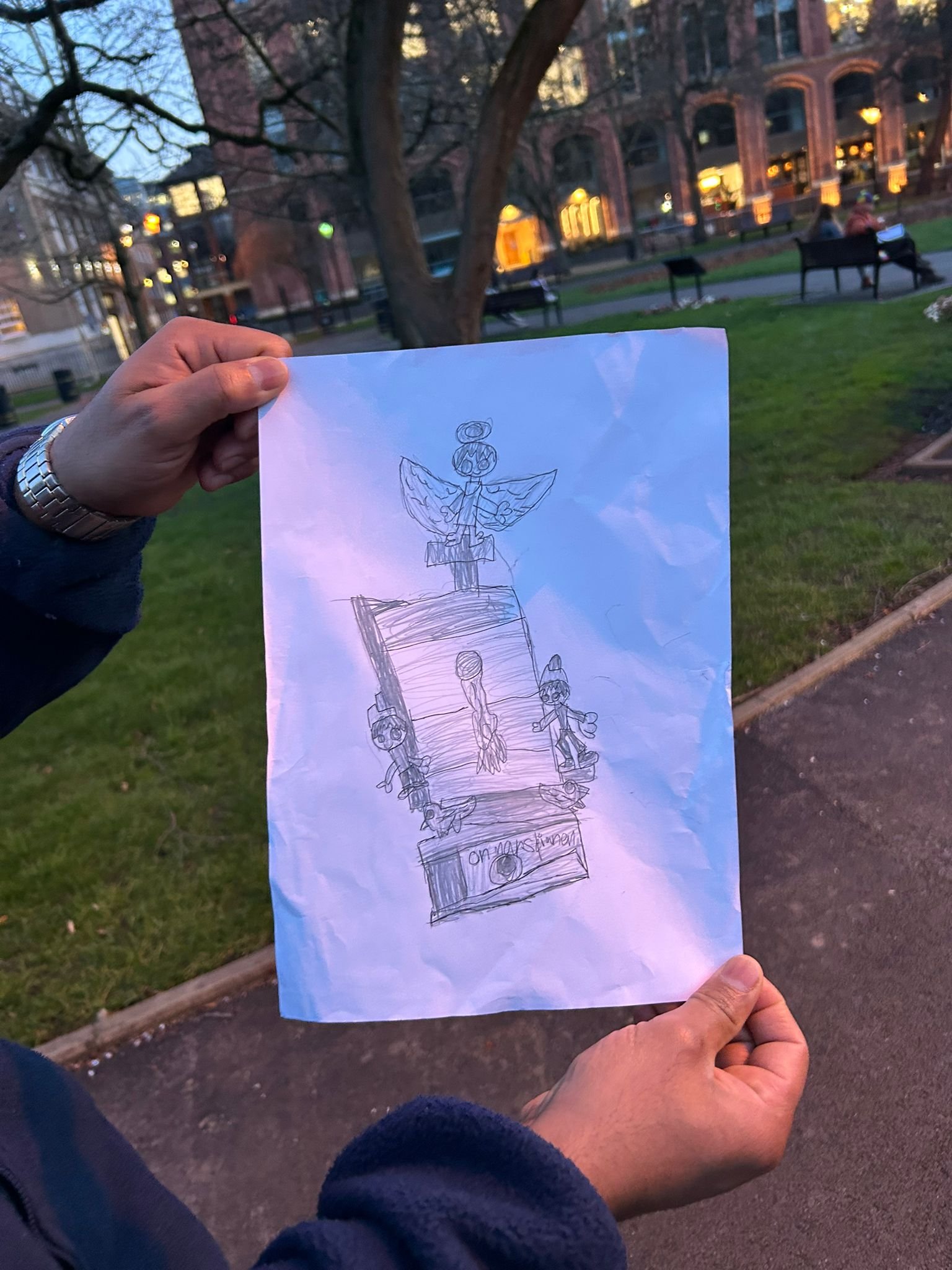 A person holding a piece of paper with a drawing of a statue with figures on top and people around it, with a park setting and benches in the background during evening or twilight.