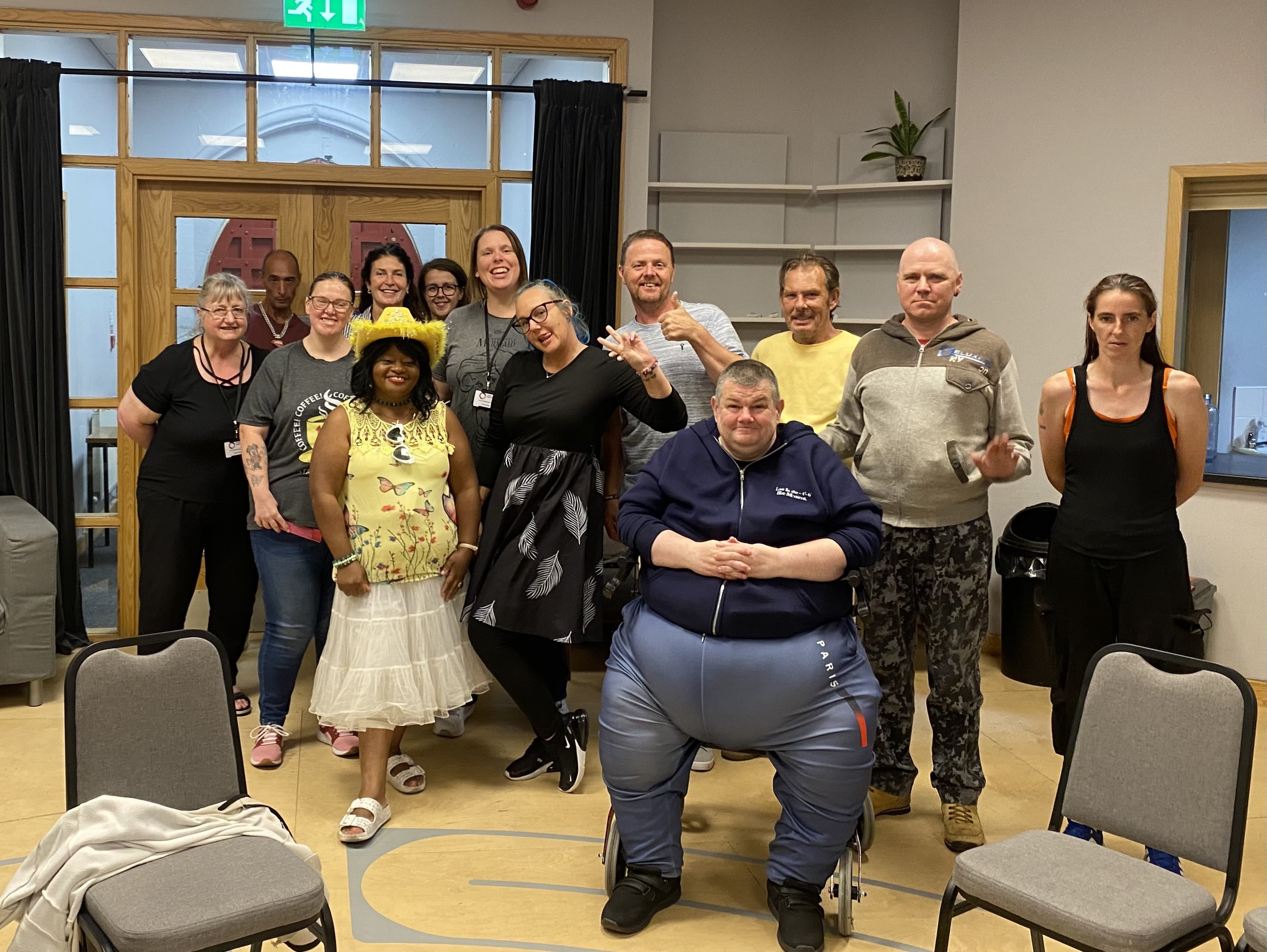 Group of people gathered indoors for a photo, some smiling and making hand gestures, in a room with wooden and glass doors, a potted plant on a shelf, and chairs around.