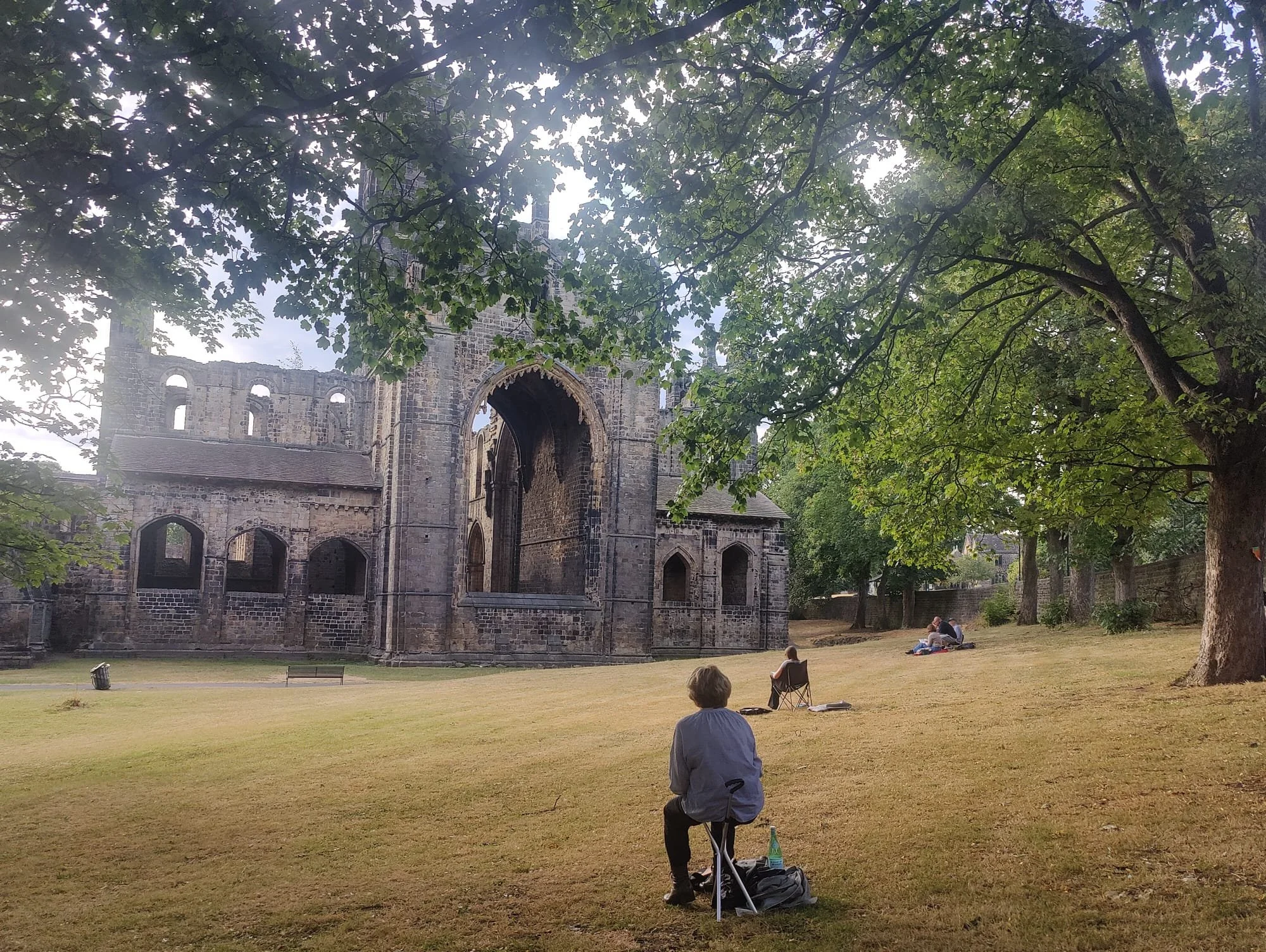 People sitting on the grass and relaxing near Kirkstall Abbey on a sunny day, surrounded by trees.