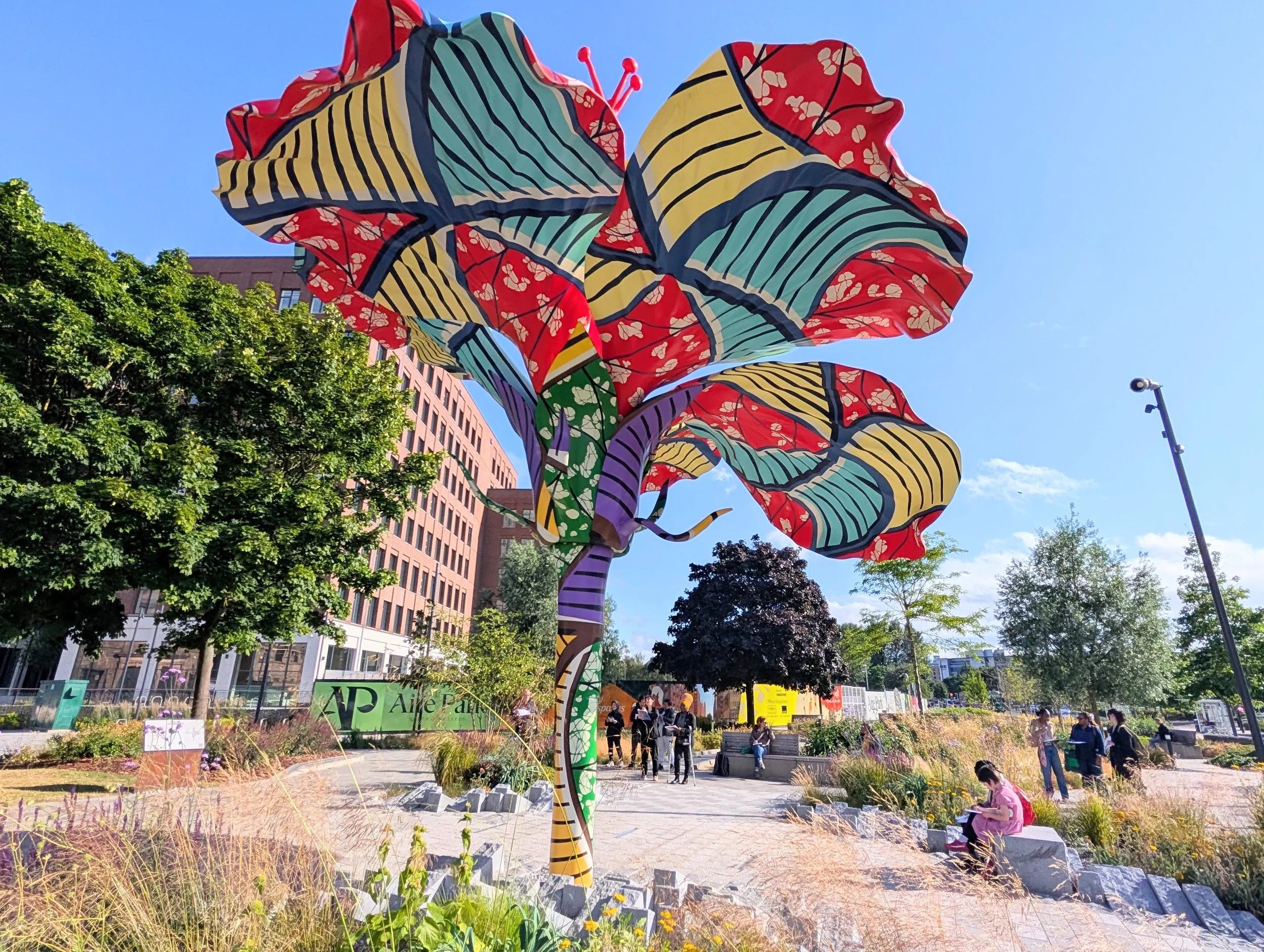 Colorful abstract sculpture resembling a flower with striped and floral patterns in red, yellow, blue, green, and purple, set in an urban park with trees, benches, and people walking and sitting.