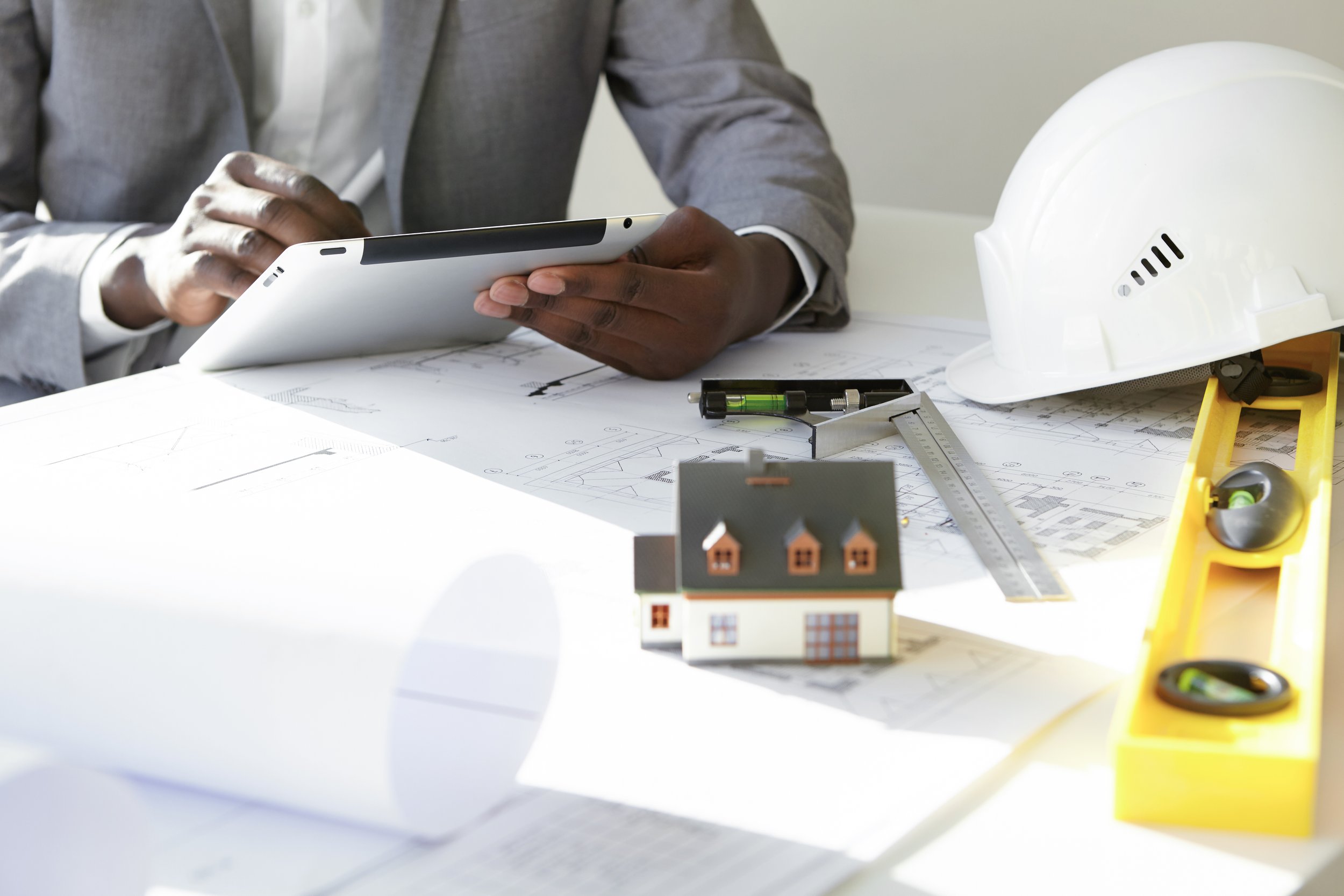cropped-shot-dark-skinned-contractor-holding-touch-pad-entering-data-while-working-new-housing-project-sitting-desk-with-drawings-scale-model-house-blueprint-rolls-ruler-helmets.jpg