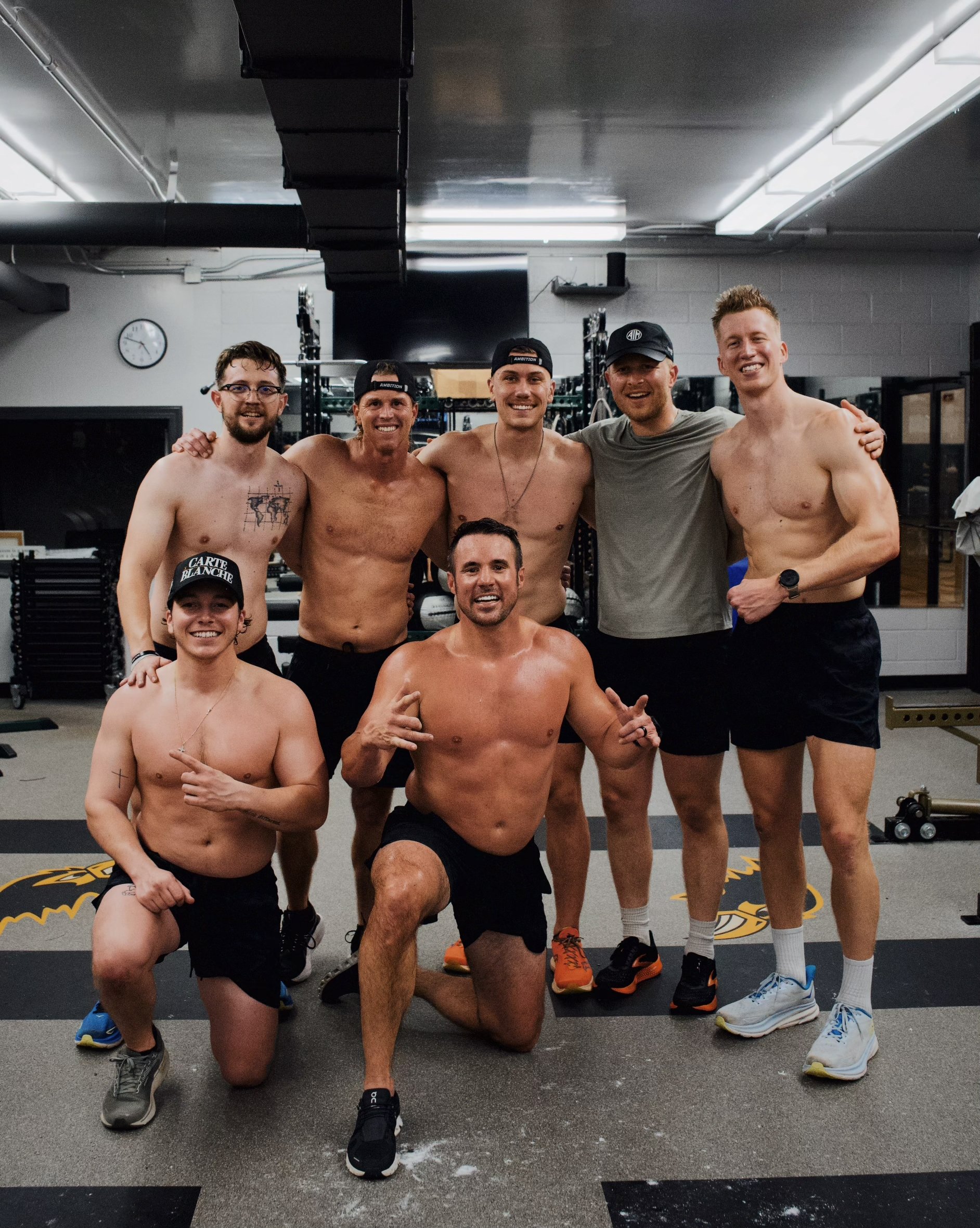 Group of seven men, some shirtless, posing in a gym setting. They are smiling and appear to be in a fitness environment with gym equipment visible. The lighting is bright and the atmosphere is casual.