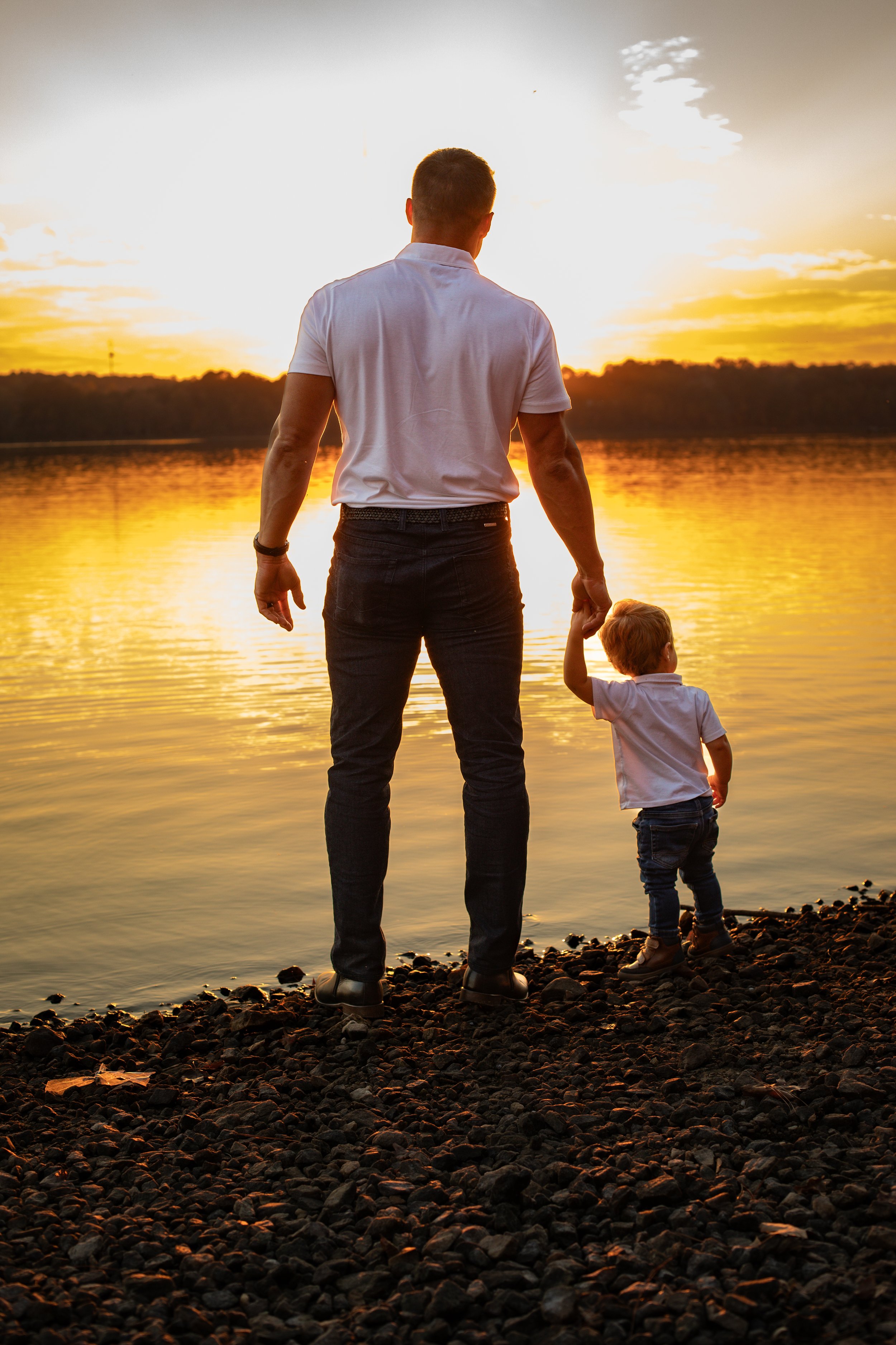 Man and child holding hands by a lake at sunset.