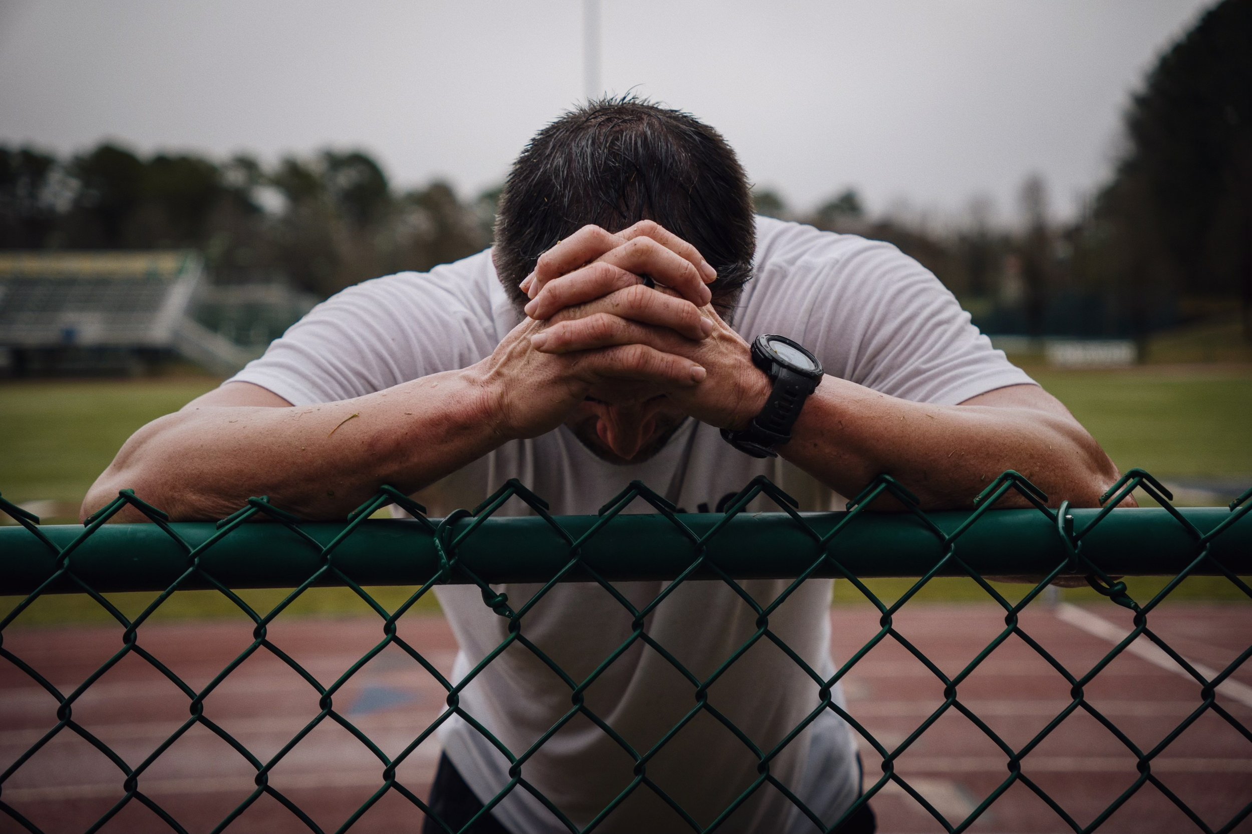A person in a white t-shirt leaning on a green chain-link fence at a sports field, head down and hands clasped together, wearing a watch.