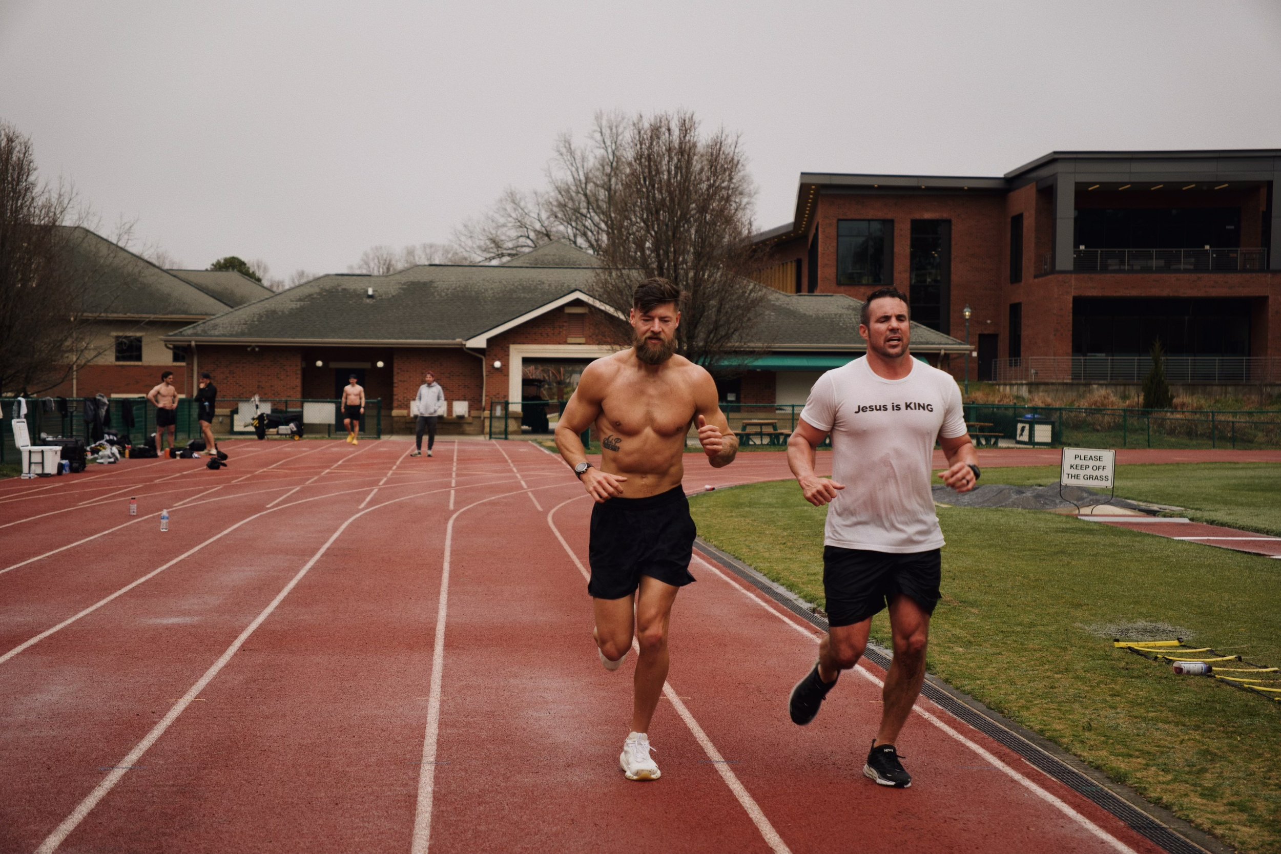 Two men jogging on a track outside, with one shirtless and the other wearing a T-shirt that reads "Jesus is King." Other people are visible in the background near a building.