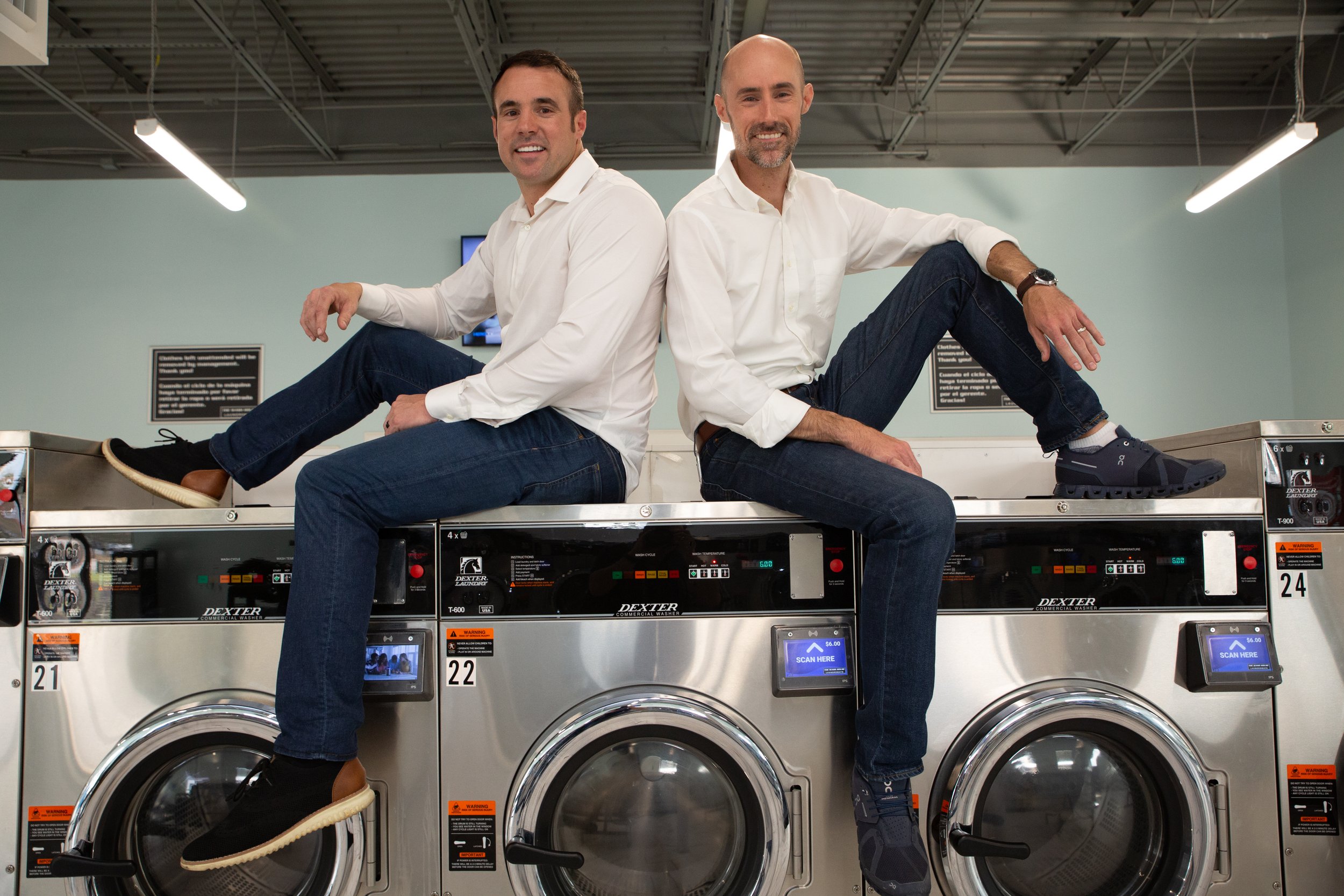 Two men in white shirts and jeans sitting on top of industrial washing machines in a laundromat.