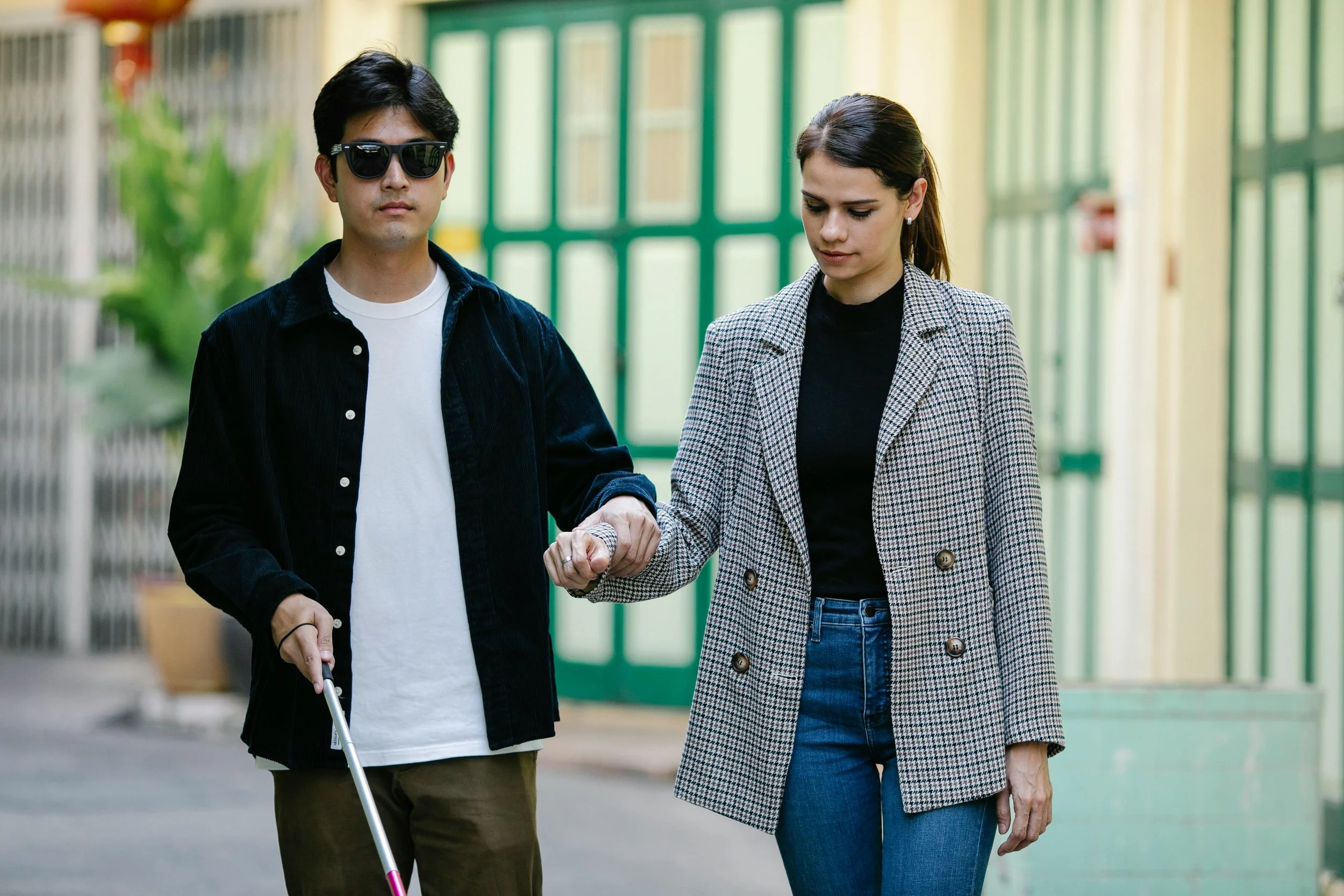 A visually impaired man wearing sunglasses and holding a white cane is guided by a woman holding his hand, walking outdoors together.