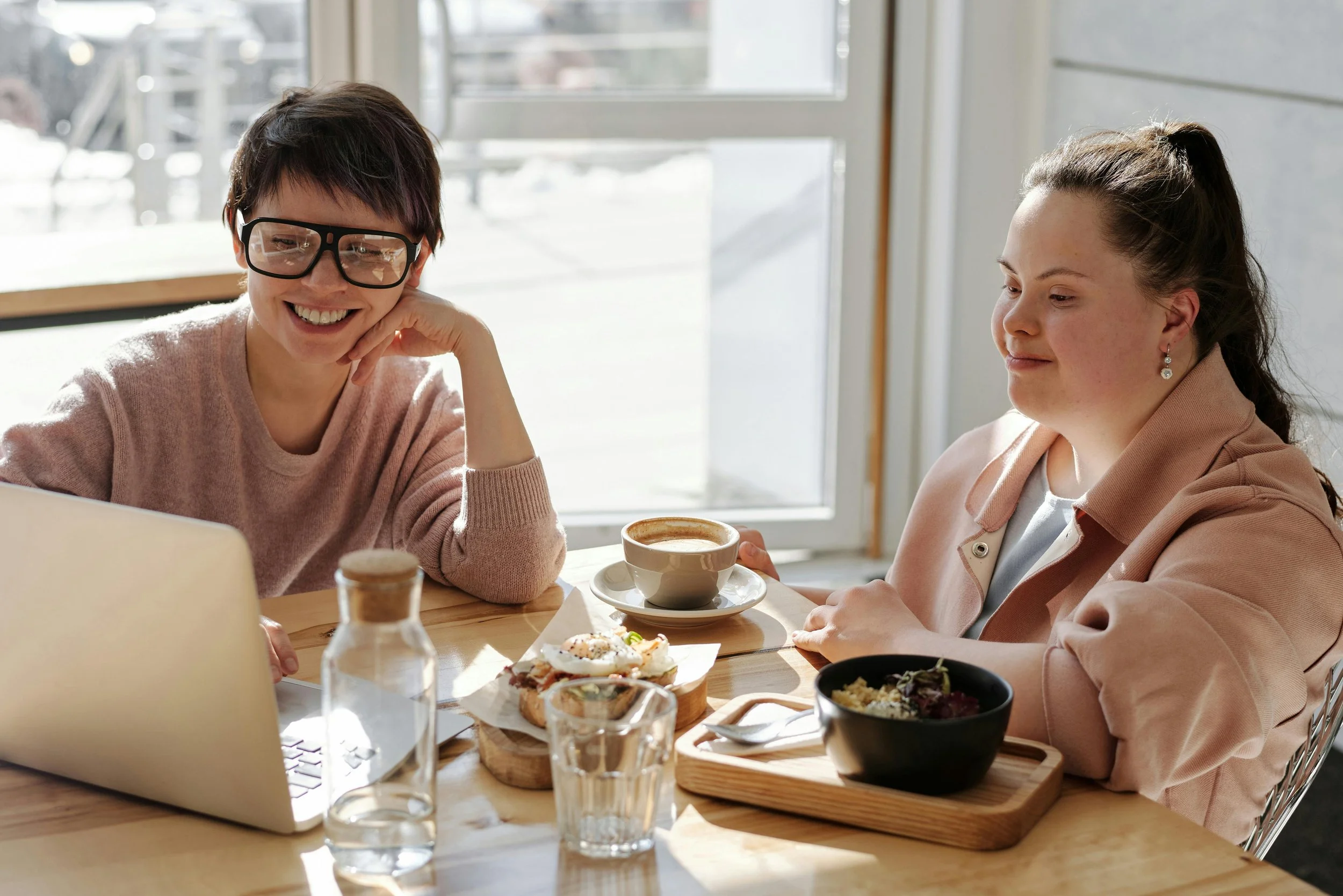 Two women sitting at a table in a coffee shop, enjoying drinks and snacks, with a laptop open in front of them, sunlight streaming through a large window behind them.